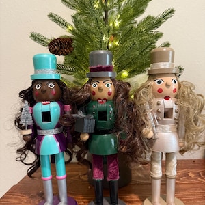 May include: Three decorative nutcrackers with long hair and top hats stand in front of a small Christmas tree. The nutcrackers are painted in teal, green, and beige with matching bases. Each holds a small accessory.