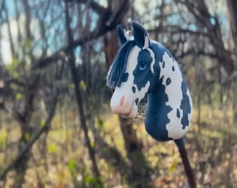 May include: A black and white spotted hobby horse with a blue mane and a blue eye. The horse is standing in a wooded area.