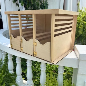 May include: A light-colored wooden storage organizer featuring three individual compartments. Each section has a hinged door with gold-tone metal hardware and a scalloped top edge. The upper portion of each compartment includes horizontal slats.
