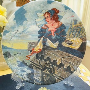 Puede incluir: Plato decorativo con una pintura de una mujer con un vestido azul sobre un muro de piedra con vistas al mar. El plato se exhibe sobre un soporte transparente, con un mantel amarillo y flores en el fondo.