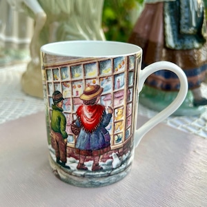 Può includere: Una tazza in ceramica bianca con un'illustrazione colorata di due bambini che guardano la vetrina di un negozio di giocattoli. La vetrina è piena di giocattoli e i bambini indossano vestiti invernali. La tazza ha un manico ed è decorata con un tema natalizio festivo.