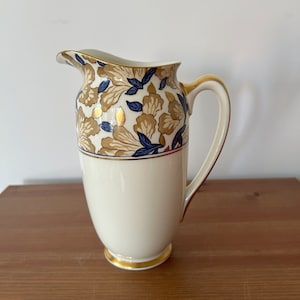 May include: A white ceramic pitcher with a gold and blue floral design. The pitcher has a wide mouth and a handle.
