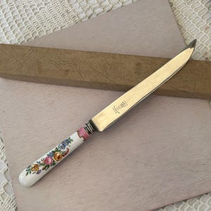 May include: A silver butter knife with a floral patterned handle. The handle is white with pink, yellow, and blue flowers. The knife is in a brown box.