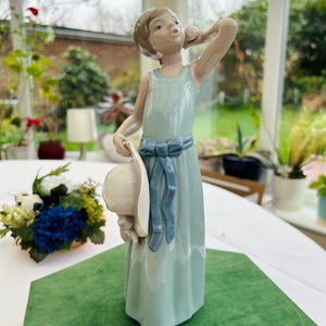 May include: A porcelain figurine of a girl in a light blue dress holding a white hat. She is standing on a green base and has a blue ribbon around her waist.