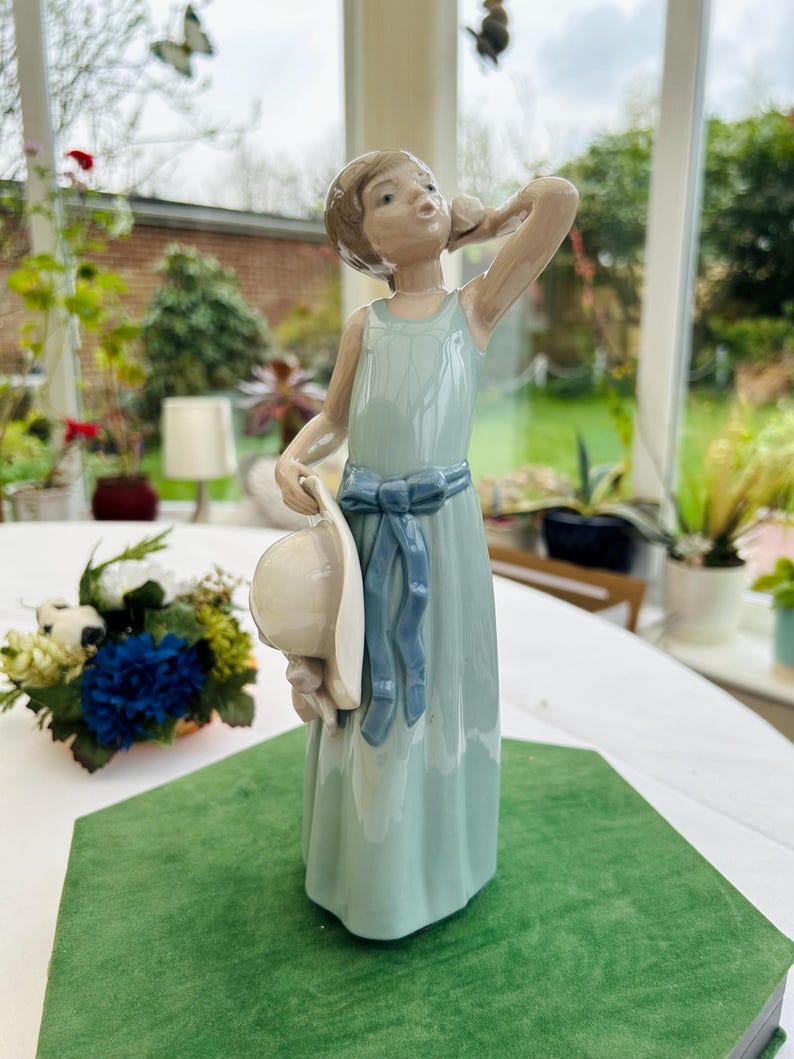 May include: A porcelain figurine of a girl in a light blue dress holding a white hat. She is standing on a green base and has a blue ribbon around her waist.