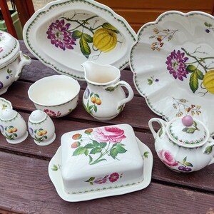 Puede incluir: Un juego de platos de porcelana blanca con diseños florales y de frutas. El juego incluye un tazón grande para servir, un tazón más pequeño, una jarra de crema, una mantequillera, un azucarero, dos saleros y pimenteros y un plato ovalado pequeño.