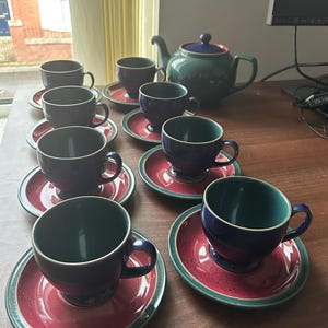 May include: A tea set consisting of a teapot and seven matching cups and saucers. Each piece features a deep blue and teal exterior with a speckled red center on the saucers. The teapot has a matching blue lid and a curved handle and spout.