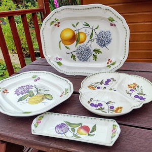 May include: Four white ceramic serving dishes with green trim and floral designs featuring fruit and flowers. The dishes are on a wooden surface.