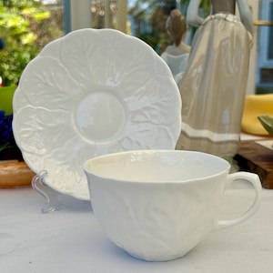 Könnte beinhalten: Eine weiße Teetasse und Untertasse mit einem Blattdesign. Tasse und Untertasse sind aus feinem Porzellan gefertigt und haben ein zartes, elegantes Aussehen.