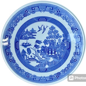 May include: A blue and white ceramic plate featuring a traditional willow pattern design. The plate depicts a central image of a pagoda, a willow tree, and a boat. The border of the plate is decorated with a repeating pattern of stylised flowers and leaves.