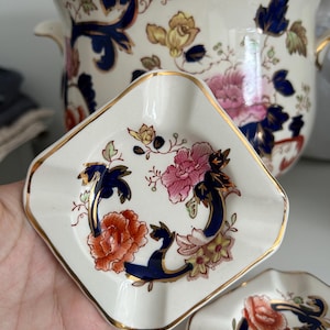 Op de afbeelding: Twee vierkante asbakken van keramiek met een blauw en gouden bloemenmotief. De asbakken hebben een gegolfde rand en zijn versierd met roze, oranje en gele bloemen.