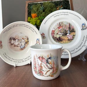 May include: A white ceramic dinnerware set featuring three pieces: a bowl, a plate, and a mug. Each piece is decorated with colorful illustrations of ducks and foxes from Beatrix Potter stories, accompanied by printed text from the tales.