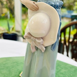 May include: A porcelain figurine of a woman wearing a blue dress and holding a large white hat with a pink ribbon.