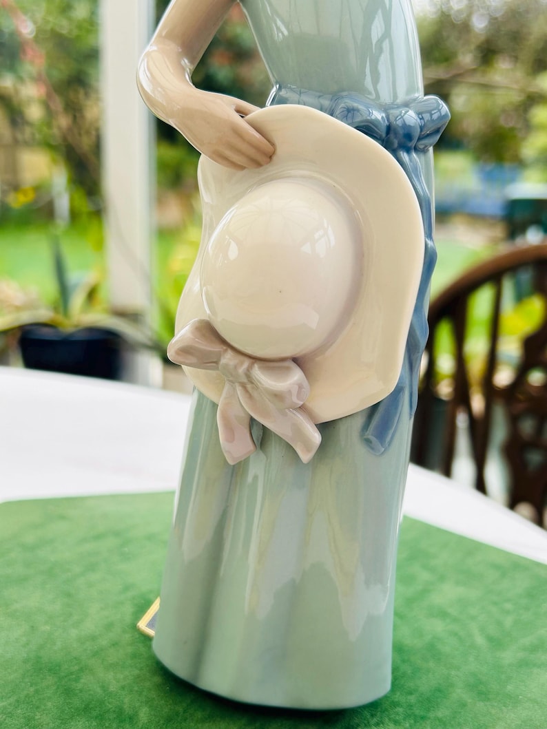 May include: A porcelain figurine of a woman wearing a blue dress and holding a large white hat with a pink ribbon.