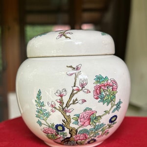 Puede incluir: Jarrón de cerámica blanca con tapa, con un diseño floral con detalles en rosa, verde y dorado. El jarrón tiene una forma redonda y un acabado liso y brillante.