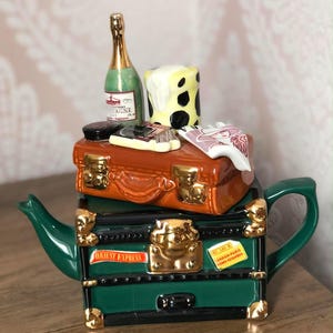 Könnte beinhalten: Eine Keramik-Teekanne in Form von gestapeltem Gepäck, grün und braun, mit goldenen Akzenten. Sie zeigt eine Champagnerflasche, einen Käseblock und andere Reiseartikel. Die Teekanne trägt die Aufschrift "Orient Express" an der Seite.