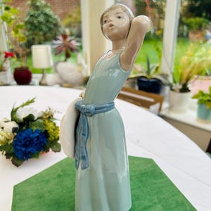 May include: A porcelain figurine of a girl in a light blue dress holding a white hat. The figurine is standing on a green base.