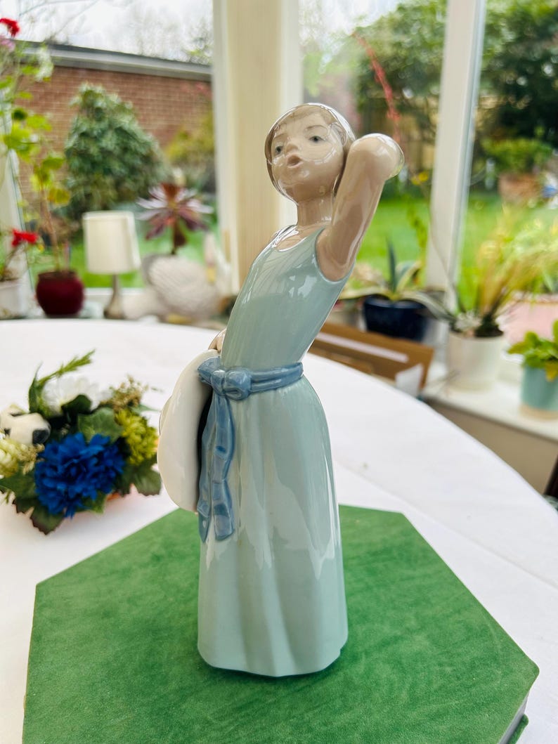 May include: A porcelain figurine of a girl in a light blue dress holding a white hat. The figurine is standing on a green base.