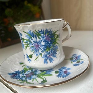 May include: White teacup and saucer set with a floral design featuring blue and purple flowers and green leaves. The cup and saucer have gold trim. The teacup is resting on the saucer.