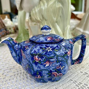 Könnte beinhalten: Eine blaue und weiße Teekanne mit Blumendekor und Deckel. Die Teekanne hat ein Blumenmuster mit rosa, weißen und blauen Blumen. Die Teekanne steht auf einer weißen Häkeltischdecke.