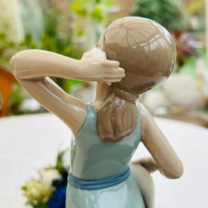 May include: A porcelain figurine of a girl wearing a light blue dress with a blue sash. She is holding a seashell to her ear and has brown hair.