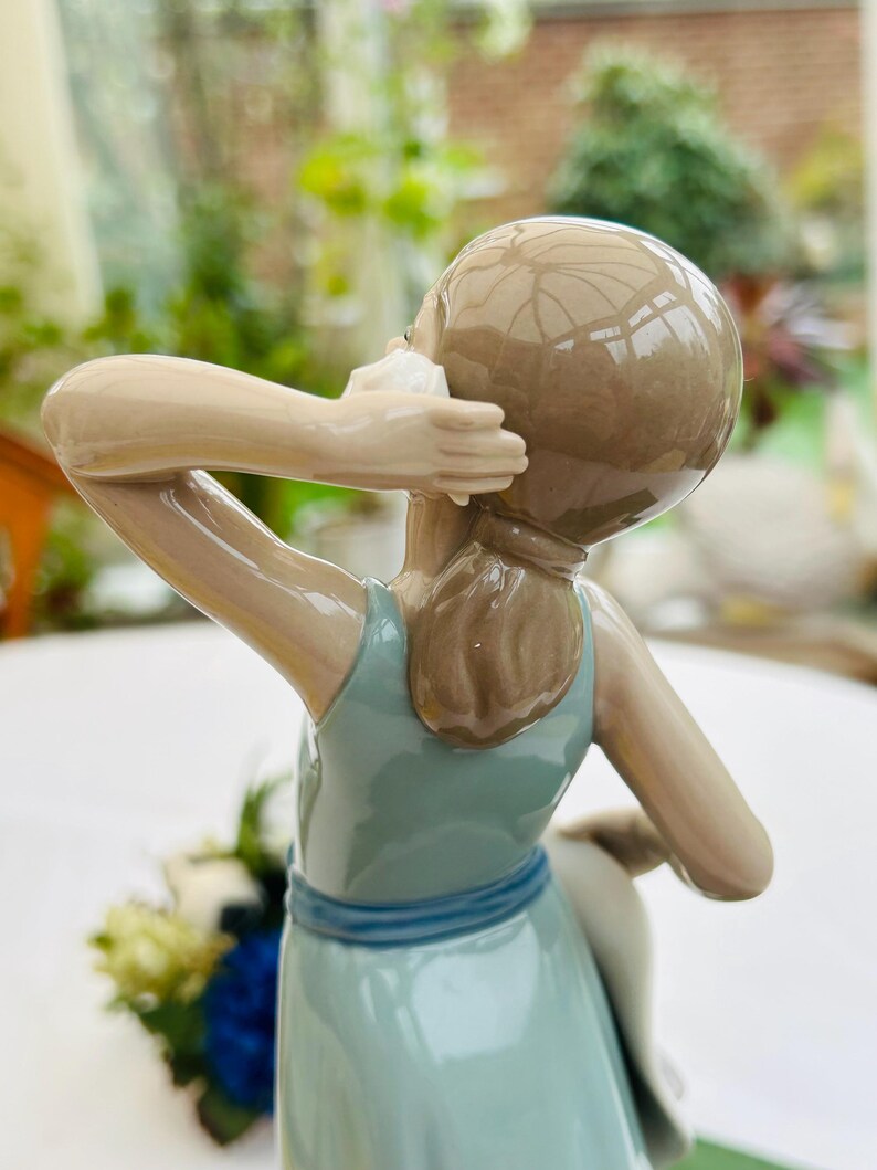 May include: A porcelain figurine of a girl wearing a light blue dress with a blue sash. She is holding a seashell to her ear and has brown hair.
