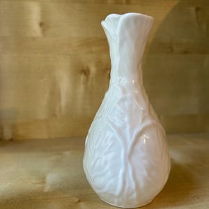 Może przedstawiać: Biały ceramiczny wazon z błyszczącym wykończeniem i teksturowaną powierzchnią z wypukłym wzorem liści, który owija się wokół korpusu. Wazon ma wąską szyjkę, która lekko rozszerza się przy górnym otworze oraz zaokrągloną, pękatą podstawę.