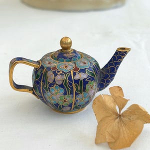 Puede incluir: Pequeña tetera azul con un diseño floral en tonos azules, verdes y morados. Cuenta con detalles dorados en el asa, el pico y la base. La tapa está adornada con un pomo dorado. Artículo decorativo.