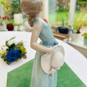 May include: A porcelain figurine of a girl in a light blue dress holding a white hat. The figurine is standing on a green surface.