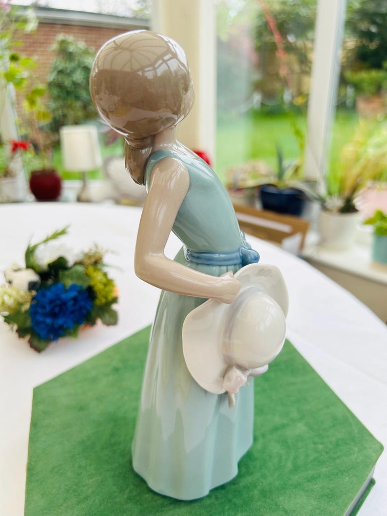 May include: A porcelain figurine of a girl in a light blue dress holding a white hat. The figurine is standing on a green surface.