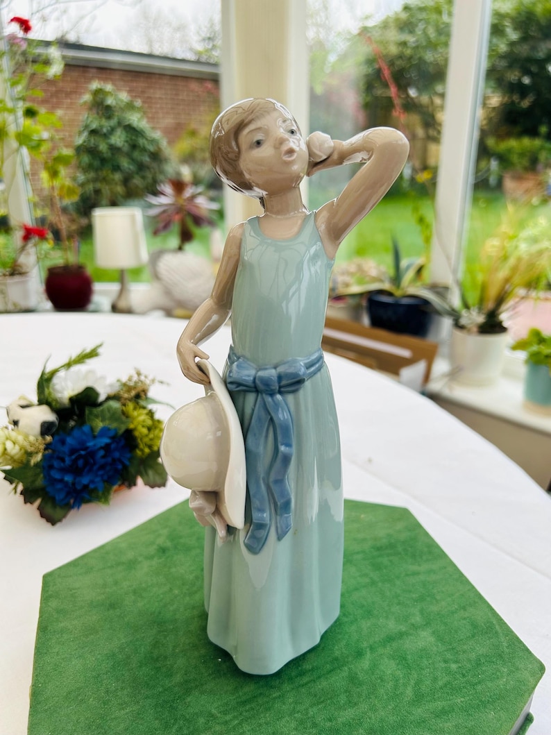 May include: A porcelain figurine of a girl wearing a light blue dress with a blue sash and holding a white hat. The figurine is standing on a green surface.