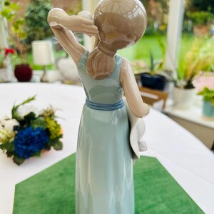 May include: A porcelain figurine of a woman in a light blue dress holding a white hat. The figurine is standing on a green surface.