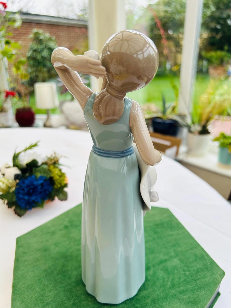 May include: A porcelain figurine of a woman in a light blue dress holding a white hat. The figurine is standing on a green surface.