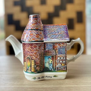 May include: A ceramic teapot shaped like a brick building with a chimney, a door, and windows. The teapot is decorated with a colorful brick pattern and a scene of a person working in a pottery shop. The text "The Old Pottery" is printed on the front of the teapot.