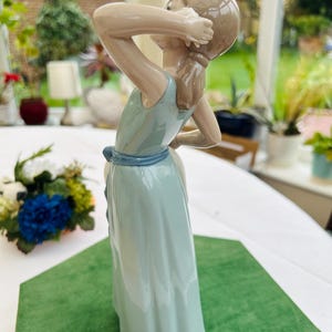 May include: A porcelain figurine of a girl in a light blue dress with a blue sash. She is holding her hair with her right hand and looking to the side. The figurine is standing on a green surface.