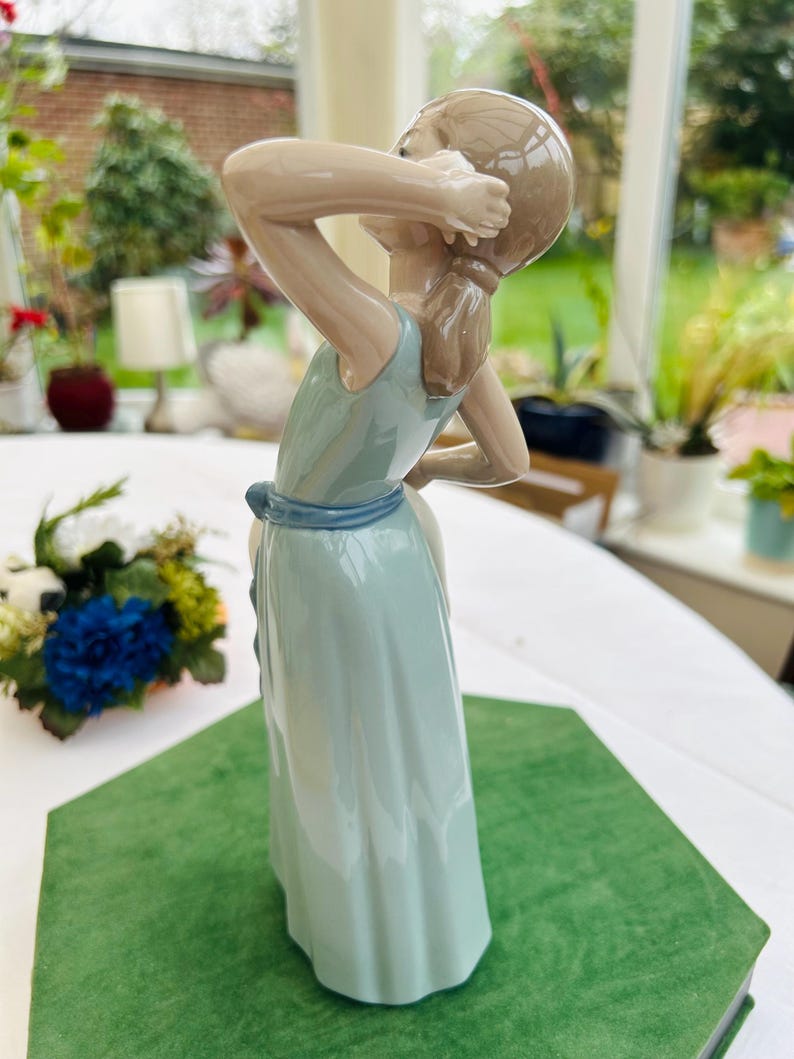 May include: A porcelain figurine of a girl in a light blue dress with a blue sash. She is holding her hair with her right hand and looking to the side. The figurine is standing on a green surface.