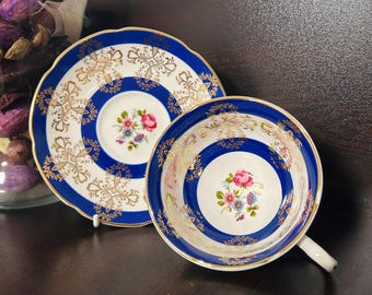 May include: A vintage teacup and saucer set. The cup and saucer have a white base with a blue and gold rim and floral centre design. The cup has a white handle. The saucer has a scalloped edge.