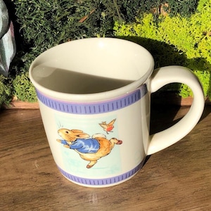 May include: White ceramic mug featuring a rabbit in a blue jacket and a bird illustration. The mug has a purple and blue striped border and a curved handle. The mug is suitable for hot beverages.