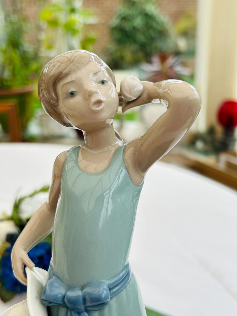 May include: A porcelain figurine of a girl wearing a blue dress and holding a white hat. She is looking to the side and has a surprised expression on her face.