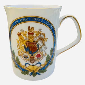 May include: White ceramic mug with a gold rim and handle. The mug features a blue and gold circular design with a coat of arms and the text "Wedding of H.R.H. Prince Charles to Lady Diana Spencer".