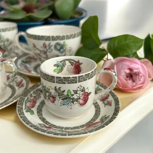 May include: A set of vintage teacups and saucers with floral patterns in red, blue, and green. The teacups have a decorative handle and sit on a matching saucer. The teacups and saucers are white with a grey border.