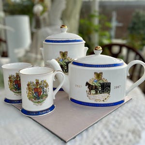 May include: A white porcelain tea set featuring a teapot, sugar bowl, and two mugs. Each piece has a blue band with gold trim and a crest design. The teapot has a horse-drawn cart illustration and the text "RINGTONS CENTENARY".