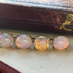Könnte beinhalten: Eine goldfarbene Brosche mit vier ovalen Opalen. Die Opale zeigen ein Farbspiel mit schillernden Farbtönen von Rosa, Blau und Gelb. Die Brosche ist vor einem neutralen Hintergrund platziert, was auf ein Schmuck- oder Antiquitätenthema hindeutet.