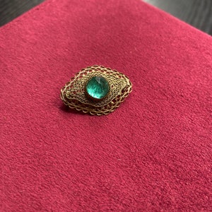 Op de afbeelding: Een vintage broche met een filigraan goudkleurig metalen frame. De broche heeft een ovale vorm en een centrale, facetgeslepen, smaragdgroene edelsteen. De broche ligt op een dieprode, getextureerde ondergrond.
