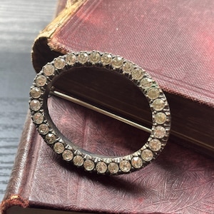 May include: An oval-shaped brooch with a silver-tone metal frame, encrusted with small, clear gemstones. The brooch has a pin on the back for attachment. The background is a vintage book with a burgundy cover.