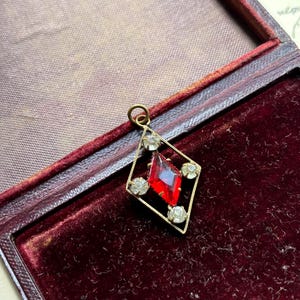 Puede incluir: Un colgante de piedra preciosa de vidrio rojo en un marco de diamante dorado con diamantes de imitación transparentes.