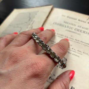 Puede incluir: Un anillo de plata con una fila de piedras transparentes facetadas. El anillo está en un dedo y el fondo es un libro.