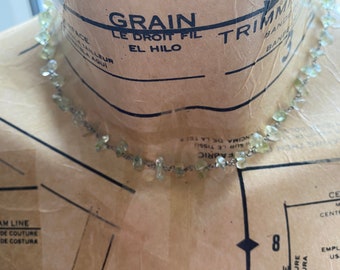May include: A light green gemstone necklace with a silver chain. The necklace is on a brown fabric background with the words "SEAM LINE", "LIGNE DE COUTURE", "LINEA DE COSTURA", "GRAIN", "LE DROIT FIL", and "EL HILO" printed on it.