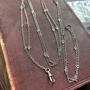 Könnte beinhalten: Eine silberfarbene Halskette mit klaren, runden Edelsteinen. Die Halskette besteht aus filigranen Ketten und hat einen Verschluss. Der Schmuck wird auf einer dunkelroten, strukturierten Oberfläche präsentiert.