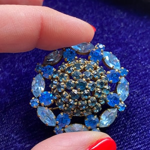 Puede incluir: Un broche vintage con un diseño circular que presenta un grupo de piedras de cristal azules y azul claro engastadas en metal dorado.
